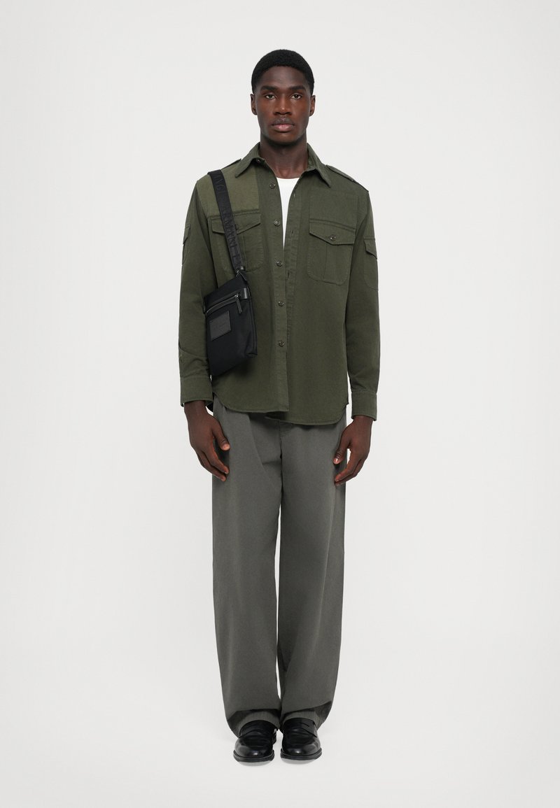 Olive green button-up shirt with two chest pockets, paired with gray wide-leg pants and black shoes. Includes a small black crossbody bag.