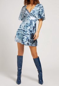 Robe à manches courtes et col en V, motif tie-dye bleu avec tissu fluide ; associée à de grandes bottes montantes en cuir verni bleu.