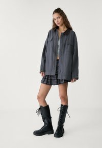 Graue übergroße Jacke mit zwei Vorderaschen, kombiniert mit einem schwarz-weißen karierten Rock. Schwarze Schnürstiefel bis zum Knie runden das Outfit ab.