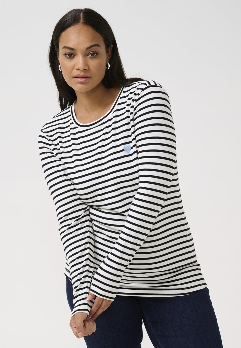 Langärmliges schwarz-weiß gestreiftes Shirt mit rundem Halsausschnitt und blauer Herzapplikation. Kombiniert mit dunklen Jeans, die enganliegende Ärmel haben.