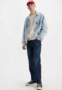 Chaqueta de mezclilla azul clara sobre un sudadera beige, combinada con jeans azul oscuro y zapatillas negras. Gorra roja con logo acentúa el look.