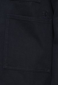 Σκοτεινός ύφασμα σε κοντινό πλάνο, δείχνοντας μια μεγάλη τετράγωνη τσέπη με ορατές ραφές και διακριτική κεντημένη λεπτομέρεια κοντά στην άνω άκρη.