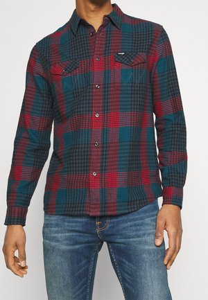 Homme portant une chemise à carreaux rouge, bleu sarcelle et noir à manches longues boutonnée avec deux poches poitrine, et un jean bleu.