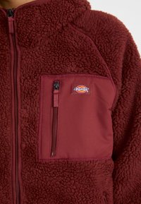 Jachetă din fleece burgund cu o textură pufoasă, având un buzunar cu fermoar pe piept și o secțiune superioară dintr-un material contrastant.