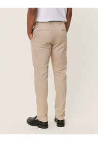 Pantalon beige en tissu lisse, coupe slim, avec deux poches arrière et une jambe légèrement effilée, porté avec des chaussures noires.