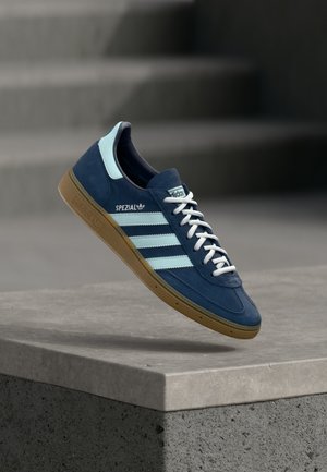 Modri čevlji Adidas Spezial s svetlo modrimi črtami in gumijastim podplatom, lebdeči nad betonsko površino z zamegljenimi stopnicami v ozadju.