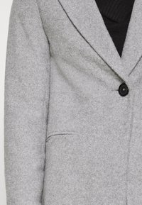 Lichtgrijze wollen blazer met een ingesneden revers, enkel zwart knoopsluiting en een voorzak. Soepele textuur met een getailleerde pasvorm.