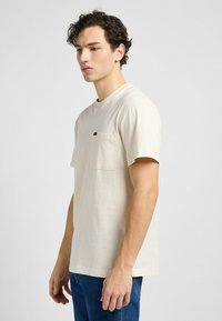 Kremowa bawełniana koszulka z krótkim rękawem, okrągłym dekoltem i kieszonką na lewej piersi. Prosty wzór bez nadruków. Model nosi niebieskie dżinsy.