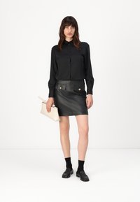 Schwarzes Hemd mit Knöpfen, kombiniert mit einem schwarzen Lederrock mit frontalen Taschen. Die Accessoires umfassen eine hellbeige Clutch und schwarze Slipper.