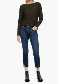Top a maniche lunghe in maglia a nido d'ape di un verde scuro abbinato a jeans skinny strappati di un blu medio e stivaletti neri alla caviglia. I jeans presentano dettagli con orlo sfrangiato.