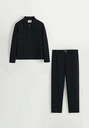 Veste noire avec un avant zippé et deux poches, assortie à un pantalon noir. Les deux pièces sont en tissu lisse et structuré.