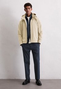 Beżowa lekka kurtka z kapturem, granatowa warstwa wewnętrzna, biały top, ciemnoszare spodnie i czarne buty. Gładka faktura, minimalistyczny design.