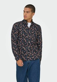 Noor mees kannab musta nööpidega särk, millel on piparkoogivaatide ja siniste lillede muster, koos siniste teksadega, vaadates kõrvale.