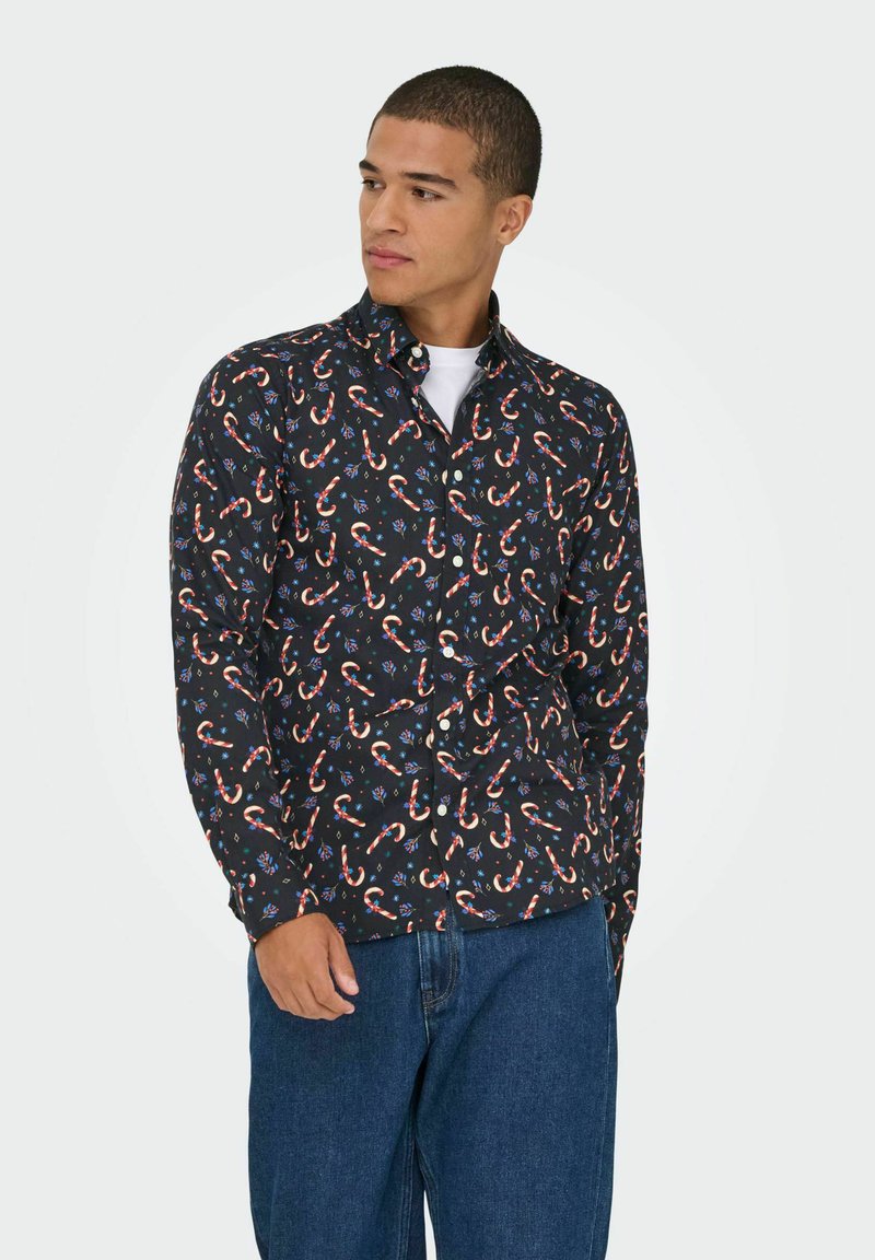 Noor mees kannab musta nööpidega särk, millel on piparkoogivaatide ja siniste lillede muster, koos siniste teksadega, vaadates kõrvale.