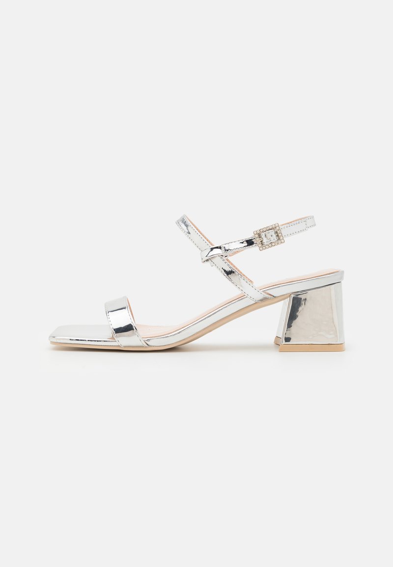 Silver patent leather sandal featuring a square toe, dual straps, and a chunky heel with a textured accent buckle.