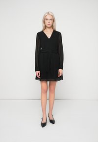 Černé šaty s dlouhými průsvitnými rukávy, výstřihem do V a páskem v pase. Lehká látka, délka ke kolenům a rozšířený lem. Kombinované s černými špičatými botami.