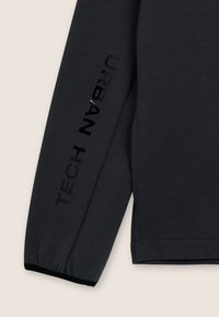 Camisa de manga larga gris oscuro con una textura acanalada. Presenta un estampado negro brillante de "TECH" y "URBAN" a lo largo de las mangas.