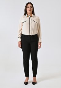 Blouse crème avec des bordures noires et deux poches poitrine, associée à un pantalon noir ajusté et des escarpins noirs pointus. Tissu lisse, coupe ajustée.