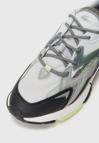 Lacoste sneakers featuring a gray mesh upper with black accents, reflective detailing, a textured toe cap, and light green hardware.