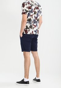 Květinová košile s krátkým rukávem na bílém podkladu s mnohobarevnými vzory, spárovaná s námořnickými šortkami. Černé boty s bílou podrážkou doplňují celý outfit.