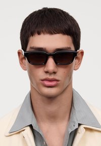 Occhiali da sole neri rettangolari con lenti scure, indossati da una persona con capelli corti, abbinati a una camicia con colletto di colore chiaro.
