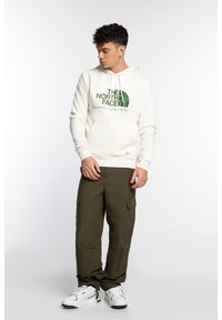 Sudadera blanca con el logo verde de "The North Face", combinada con pantalones cargo oliva y zapatillas blancas. Presenta un bolsillo tipo canguro y capucha.