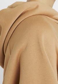 Czapka typu beanie z miękkiego beżowego materiału; ma luźne dopasowanie z opadającym kapturem i gładką fakturą, ukazując subtelne, lekkie przejścia kolorów.