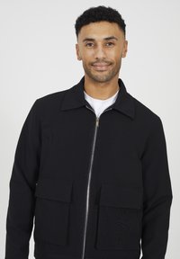 Giacca nera con zip, dalla finitura testurizzata, caratterizzata da un colletto a punta, due tasche frontali a toppa e una cerniera argentata. Design minimalista.