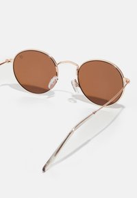 Ochelari de soare rotunzi cu lentile maro, ramă din metal auriu, suporturi pentru nas transparente și brațe subțiri. Prezintă un design minimalist.