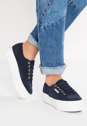 Osoba nosząca podwinięte niebieskie dżinsy i granatowe sneakersy na platformie z białymi podeszwami i złotymi oczkami na białym tle.