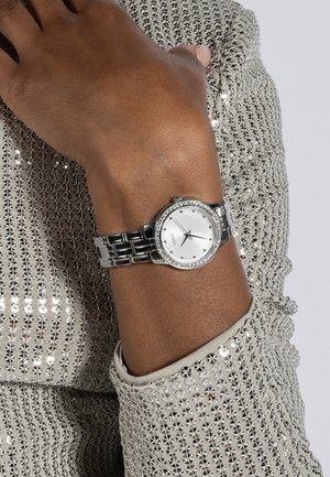 Pols met een zilveren Guess-horloge met een ronde wijzerplaat met kristalaccenten, rustend op glinsterende beige stof.