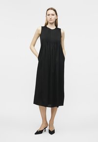 Γυναίκα που φοράει μαύρο αμάνικο μίντι φόρεμα με πλισέ μπούστο και τσέπες, φορώντας μαύρες μυτερές γόβες, μπροστά σε απλό φόντο.