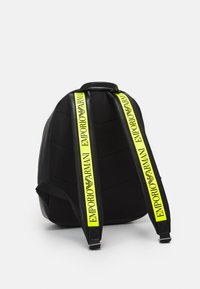 Svart ryggsäck med vadderad baksida och neongula remmar märkta "EMPORIO ARMANI" i svart text och logo mot en vit bakgrund.