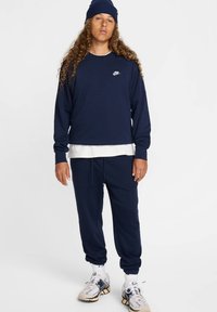 Felpa Nike blu navy a maniche lunghe e collo rotondo, indossata sopra una camicia bianca; pantaloni jogger blu navy abbinati; sneakers bianche con accenti scuri.