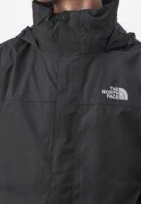Homme portant une veste noire North Face à col haut avec logo sur la poitrine et cordons sur la capuche.