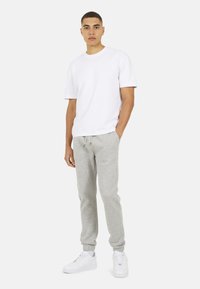 Jonge man staat, draagt een eenvoudige witte t-shirt, lichtgrijze joggingbroek en witte sneakers, tegen een neutrale achtergrond.