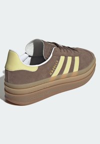 Brown suede Adidas Gazelle sneaker with yellow stripes and heel tab, thick translucent sole, and white interior lining.