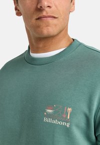 Sweatshirt vert clair avec col côtelé, arborant un logo graphique de soleil et de palmiers en couleurs corail et crème sur la poitrine.
