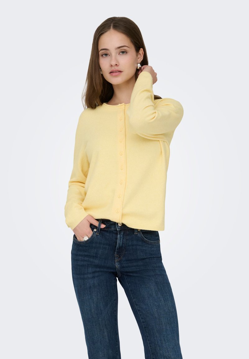 Cardigan jaune à encolure ronde, fermeture boutonnée devant et manches longues, associé à un jean en denim foncé. Texture lisse.