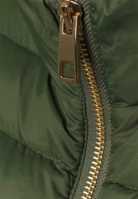 Närbild av en vadderad grön jacka med en guldkedja och en rektangulär dragflik, samt texturerad tyg som framhäver de kviltade sektionerna.