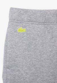 Pantalons de survêtement gris en tissu doux, arborant un logo Lacoste vert citron bien visible sur le côté gauche. Design simple avec taille élastique.