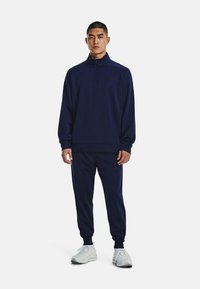 Marineblauer Trainingsanzug mit halbhohem Reißverschlusskragen und schwarzem Logo. Weicher, strukturierter Stoff. Weiße Sportschuhe. Figurbetonter Schnitt.