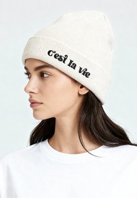 Jonge vrouw met lang bruin haar, een witte gebreide muts met de tekst "c'est la vie" en een effen wit T-shirt, kijkt naar links.