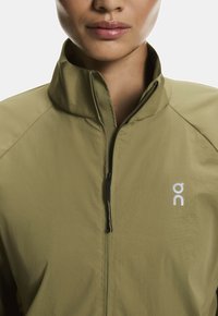 Világos olajzöld dzseki magas gallérral, elöl cipzárral és logó részlettel. Simaság, sportos dizájn és minimalista stílus.