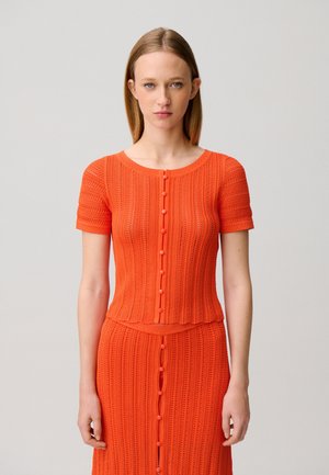 Mujer joven con cabello lacio, vestida con un top de manga corta de punto naranja brillante y una falda a juego, posando frente a un fondo liso.