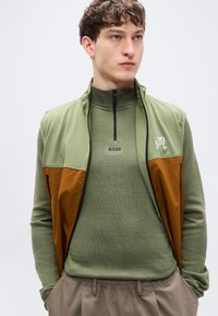 Πολυεπίπεδο γιλέκο σε πράσινο και καφέ, υφασμάτινη υφή, ψηλός γιακάς, κλείσιμο με φερμουάρ, με λογότυπο και εφαρμοστό, μακρυμάνικο τοπ από κάτω.