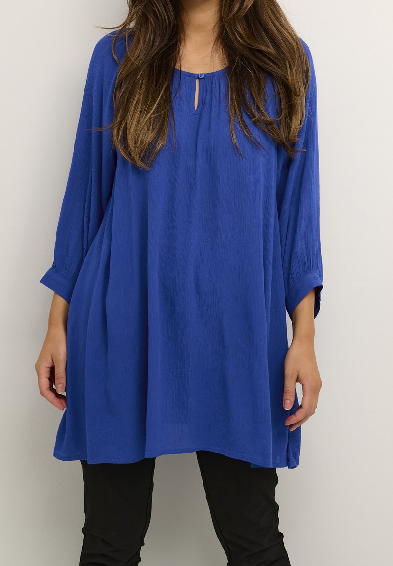Femme portant une tunique bleu roi ample à manches trois-quarts et un pantalon noir, debout devant un fond clair uni.