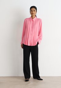 Rosa plisserte bluse med knapp-front og oversized passform, kombinert med svart vide bukser og spisse leiligheter.