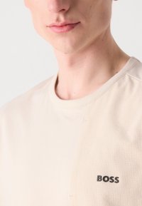 Beige, rövid ujjú póló, texturált anyagból, kontrasztos sima résszel és fekete "BOSS" logóval a bal oldalon.