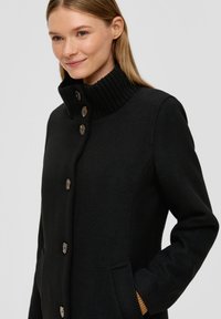 Zwarte coat met een hoge geribbelde kraag, knopen aan de voorkant en zijzakken. Gemaakt van textuur gebreide stof en heeft een getailleerde pasvorm.